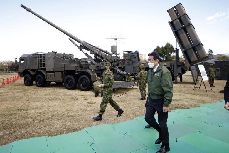 Фумио Кисида проходит мимо самоходной колесной гаубицы Type 19 калибра 155 мм (сзади слева) и ракеты класса «земля — корабль» Type 12 (сзади справа)